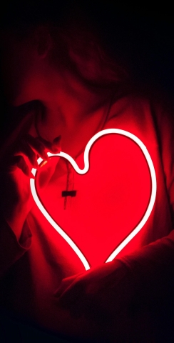 Woman in shadows holding a large, red, neon heart.