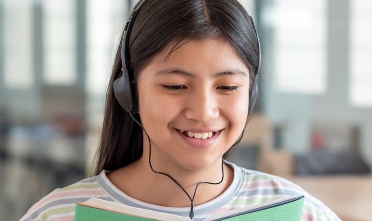 Teen girl with headphones on smiling as she looks inside a book