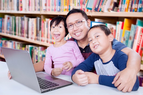 Two elementary school student and male teacher smiling at camera with laptop on the table in library