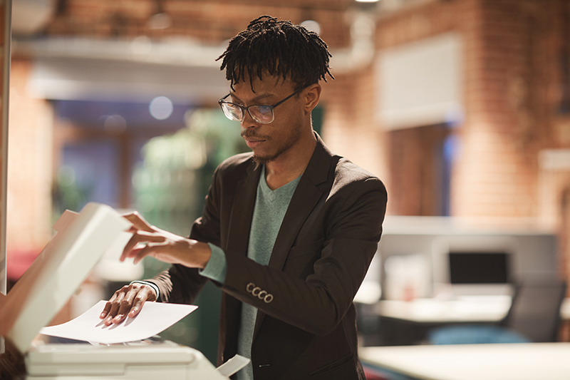 Young African businessman in eyeglasses standing near the printer and making copy of documents at office